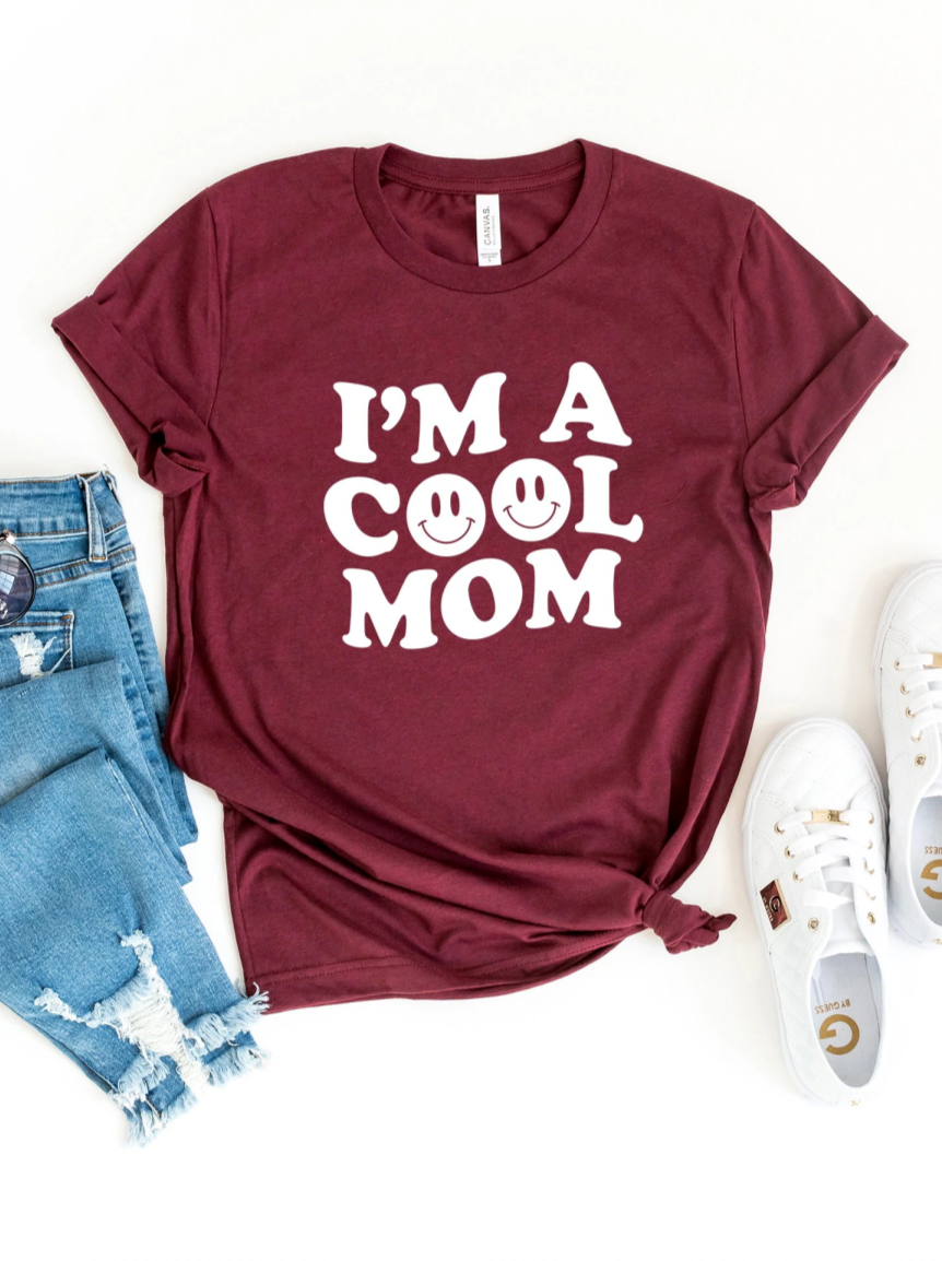 Maroon t-shirt with "I'M A COOL MOM" text and smiley faces, paired with blue jeans and white sneakers.