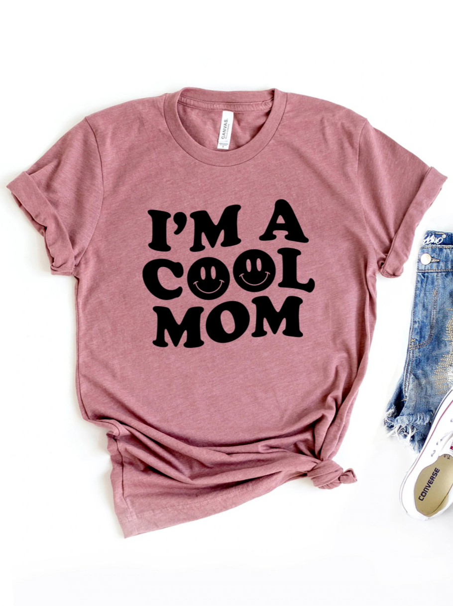 Pink t-shirt with "I'M A COOL MOM" and smiley faces printed in black. Styled with distressed jeans and sneakers.