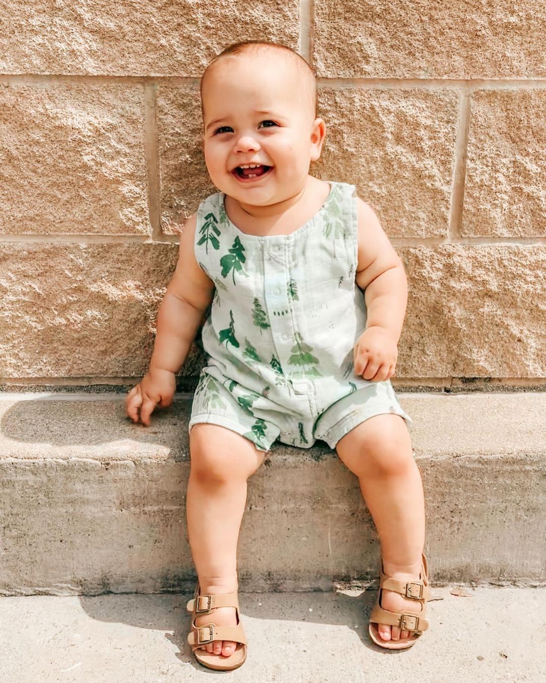 Smiling baby in green outfit sitting on a stone surface with a textured wall behind.
