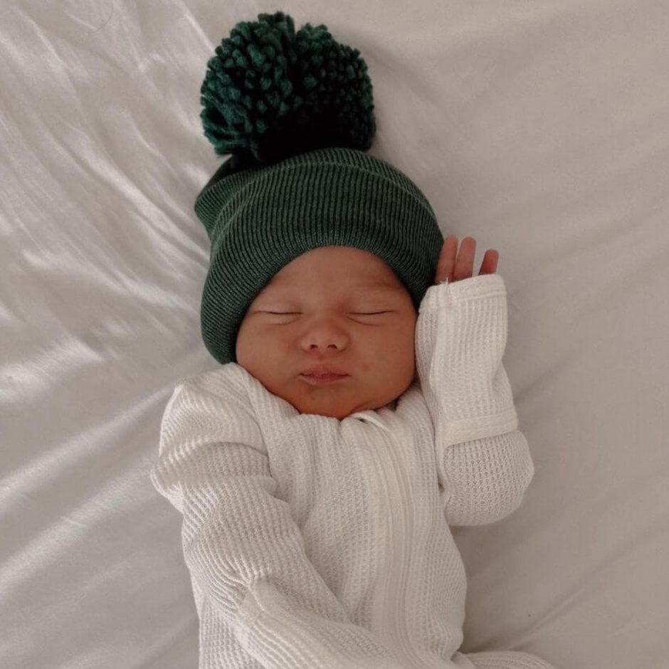 Newborn baby sleeping peacefully in a cozy green hat and white outfit on a soft white surface.
