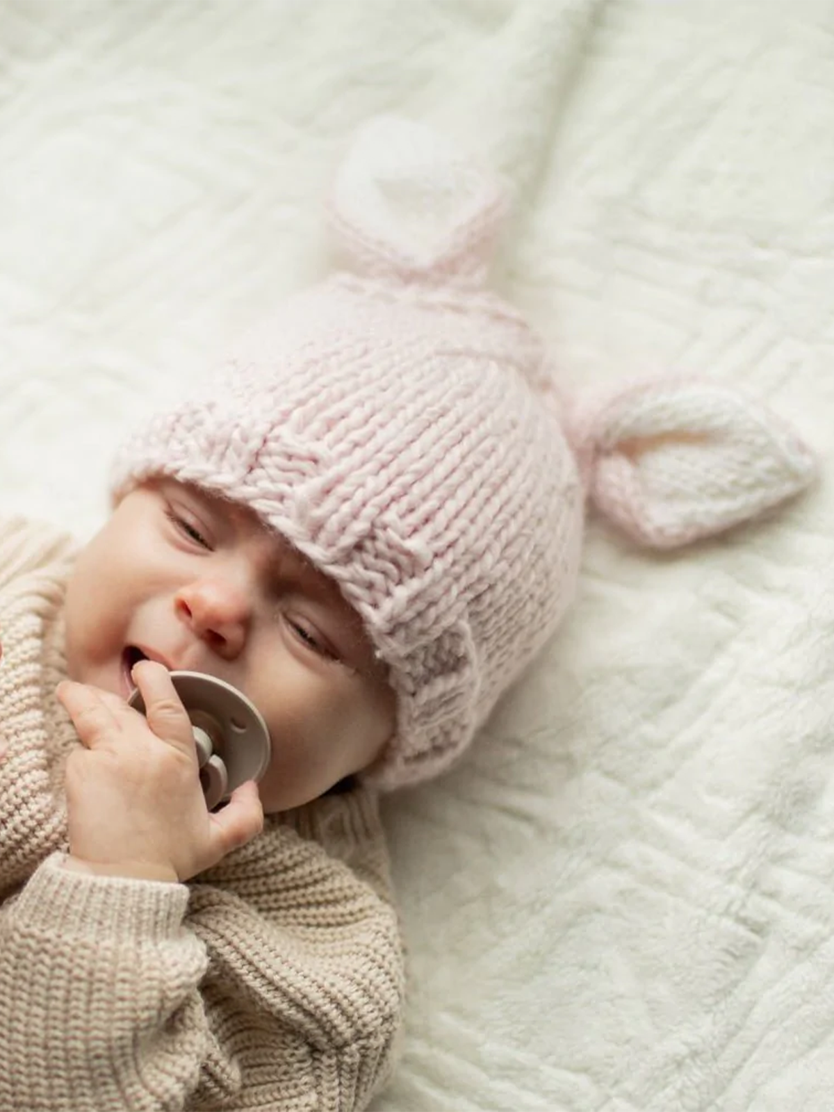 Baby wearing a knitted pink unicorn hat and beige sweater, holding a pacifier on a textured white blanket.