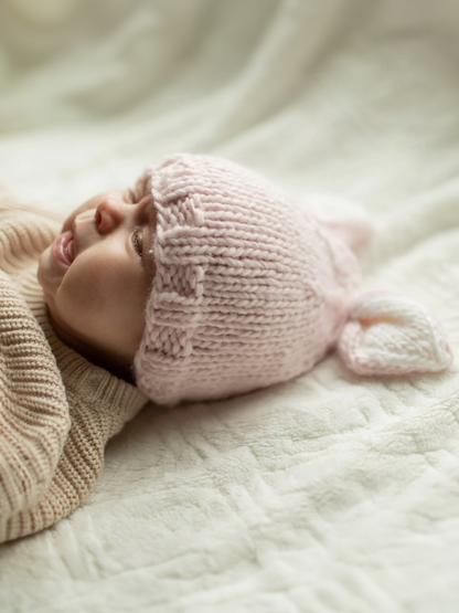 Baby in a pink knitted hat with ears, lying on a soft blanket, making a playful expression.