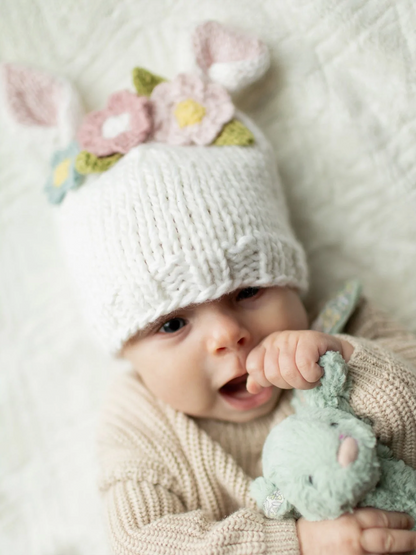 Infant in a knitted white hat with flowers and bunny ears, holding a green stuffed rabbit, smiling on a soft blanket.