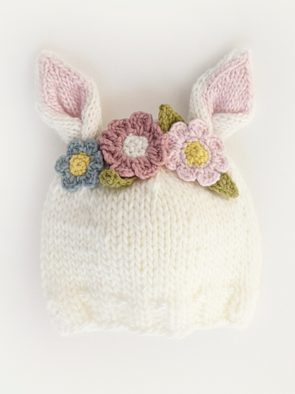 White knitted baby hat with bunny ears adorned with colorful flowers.