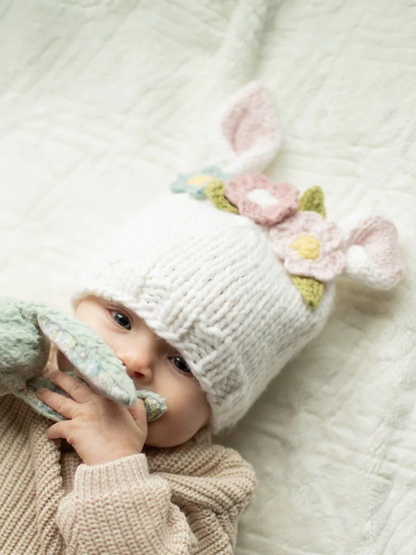 Baby wearing a knitted bunny hat with flowers, holding a soft toy, lying on a cozy blanket.