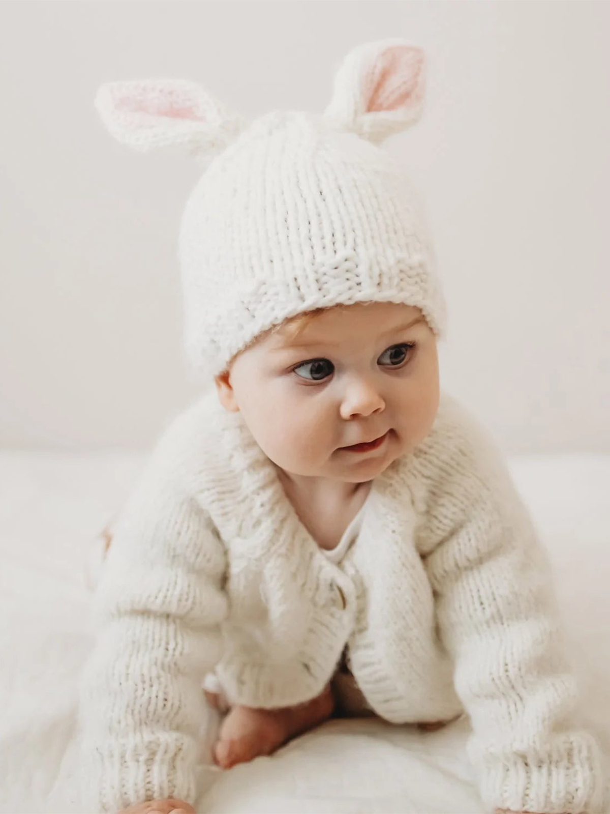 Baby in a white bunny hat and sweater sits on a white blanket, looking curiously.