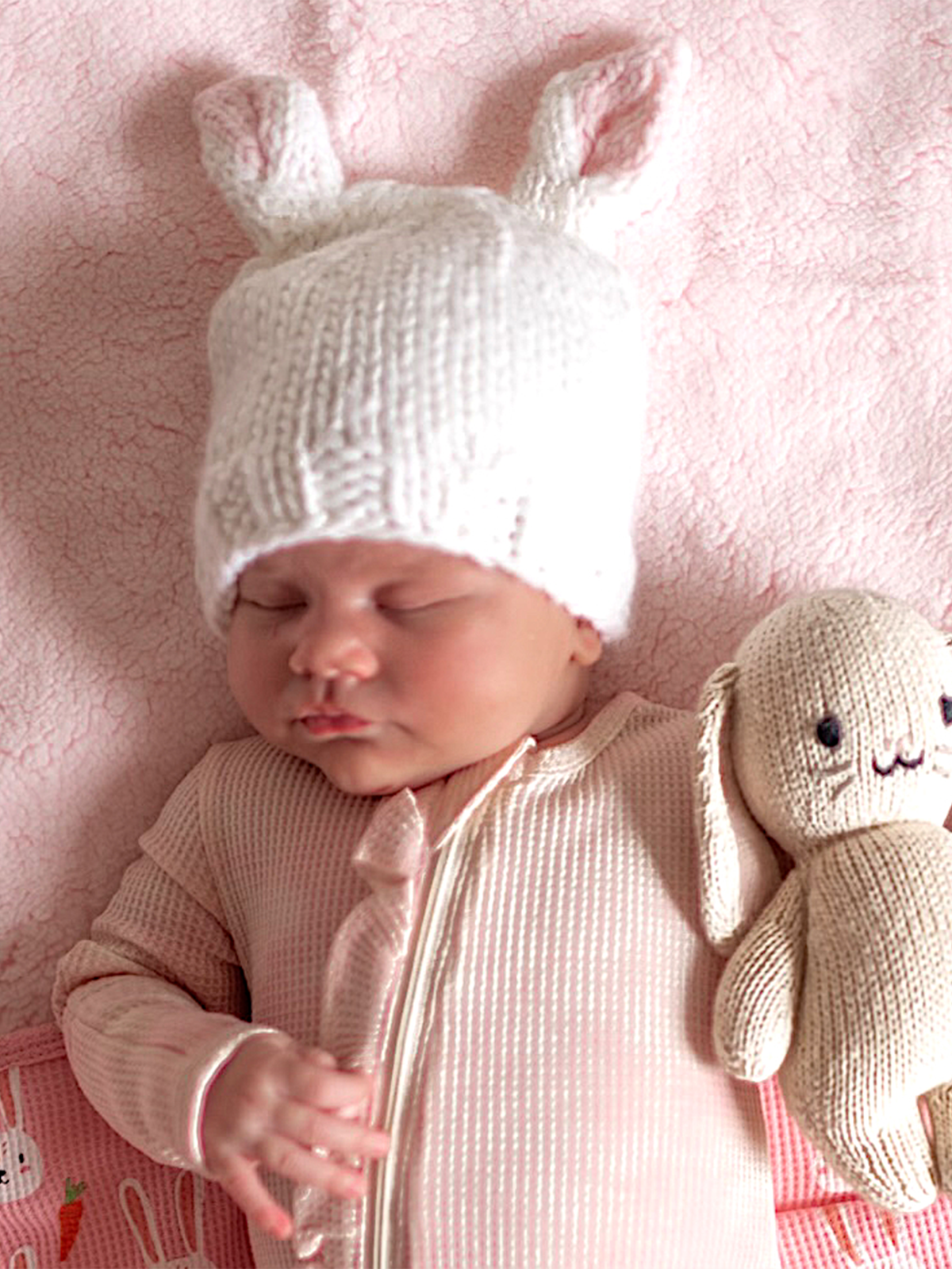 Baby in a knitted bunny hat sleeps on a pink blanket, holding a soft bunny toy.
