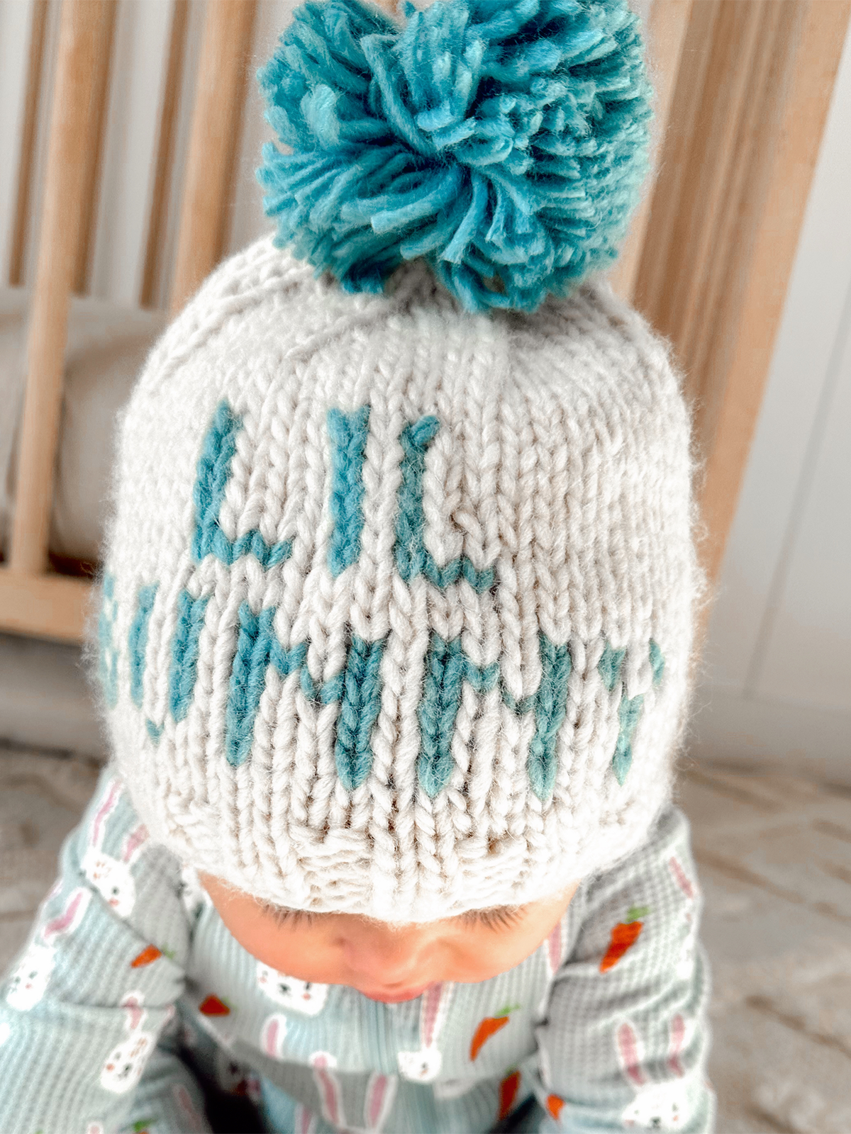 Knitted cream hat with teal pom-pom and "lil' mummy" in teal lettering, worn by a baby. Soft background and cozy setting.