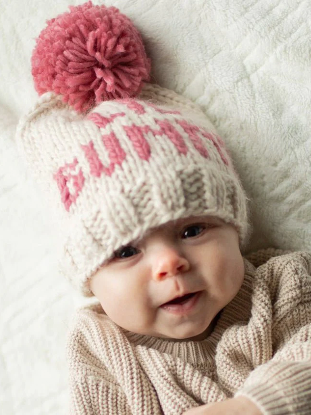 Smiling baby wearing a knitted hat with a pink pom-pom and cozy sweater, resting on a textured blanket.