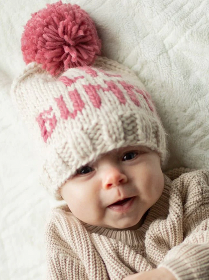 Smiling baby wearing a knitted hat with a pink pom-pom and cozy sweater, resting on a textured blanket.