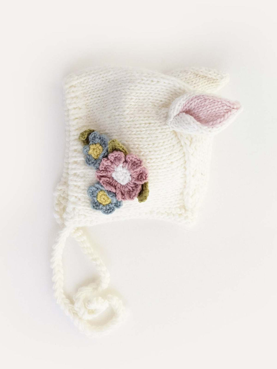 White knitted hat with floral details and bunny ears, featuring pink, blue, and green accents.