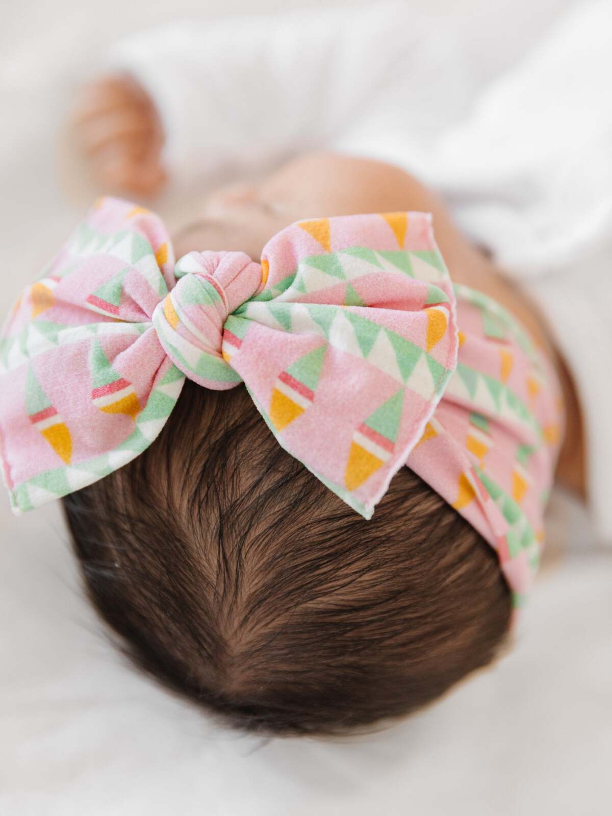 Baby girl with a colorful geometric bow headband, resting peacefully on a soft surface.