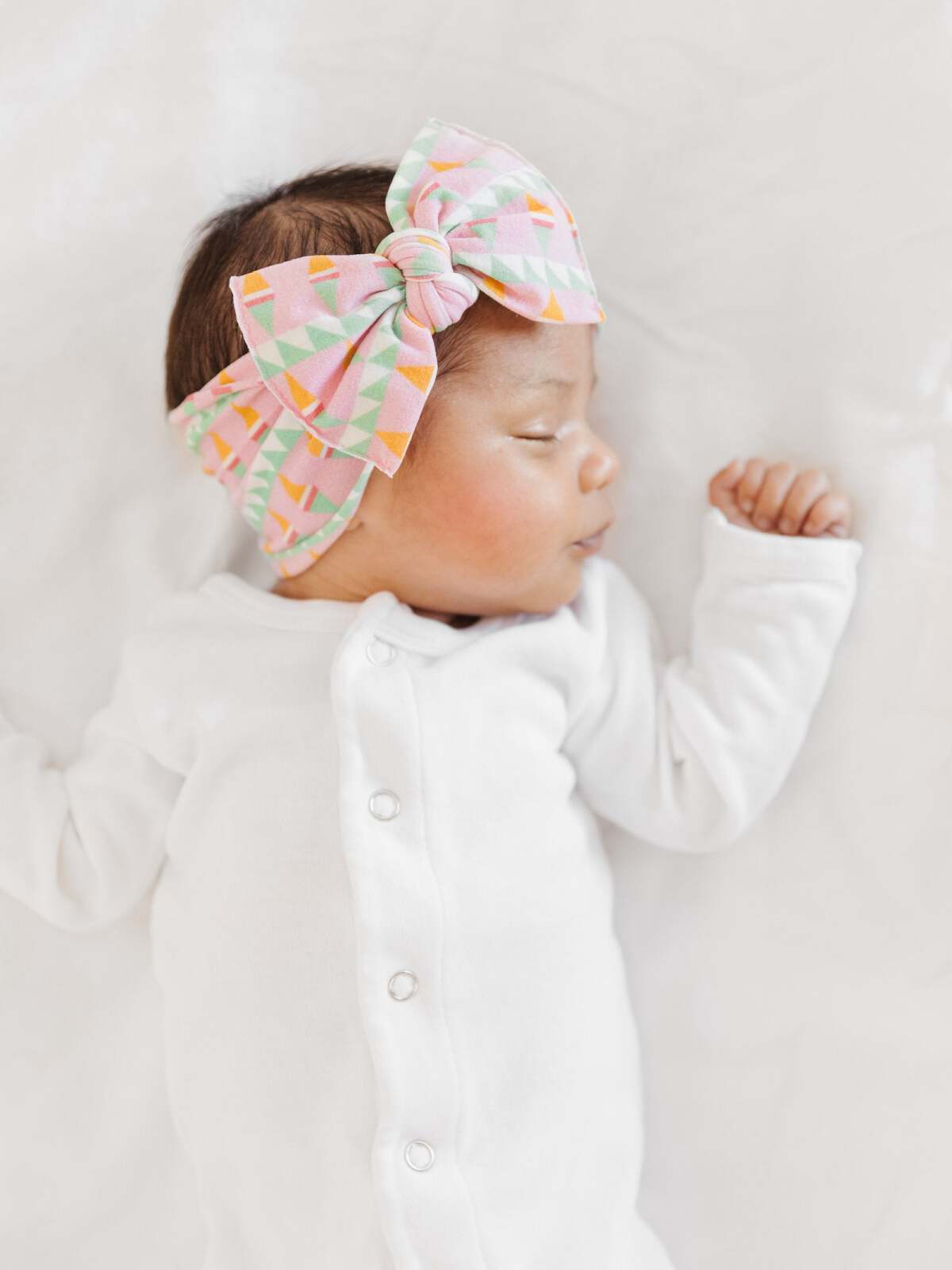 Baby girl sleeping on a soft surface, wearing a white outfit and a colorful geometric bow headband.