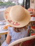 Girl wearing a wide-brimmed straw hat with "la vie est belle" embroidered, sitting at a café table.
