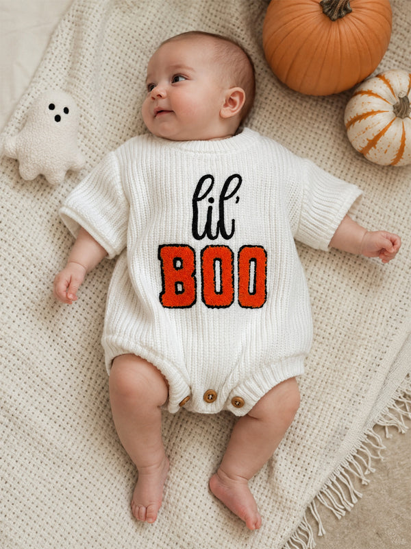 Baby in a cozy white outfit with "lil' BOO" text, surrounded by pumpkins and a ghost toy on a textured blanket.