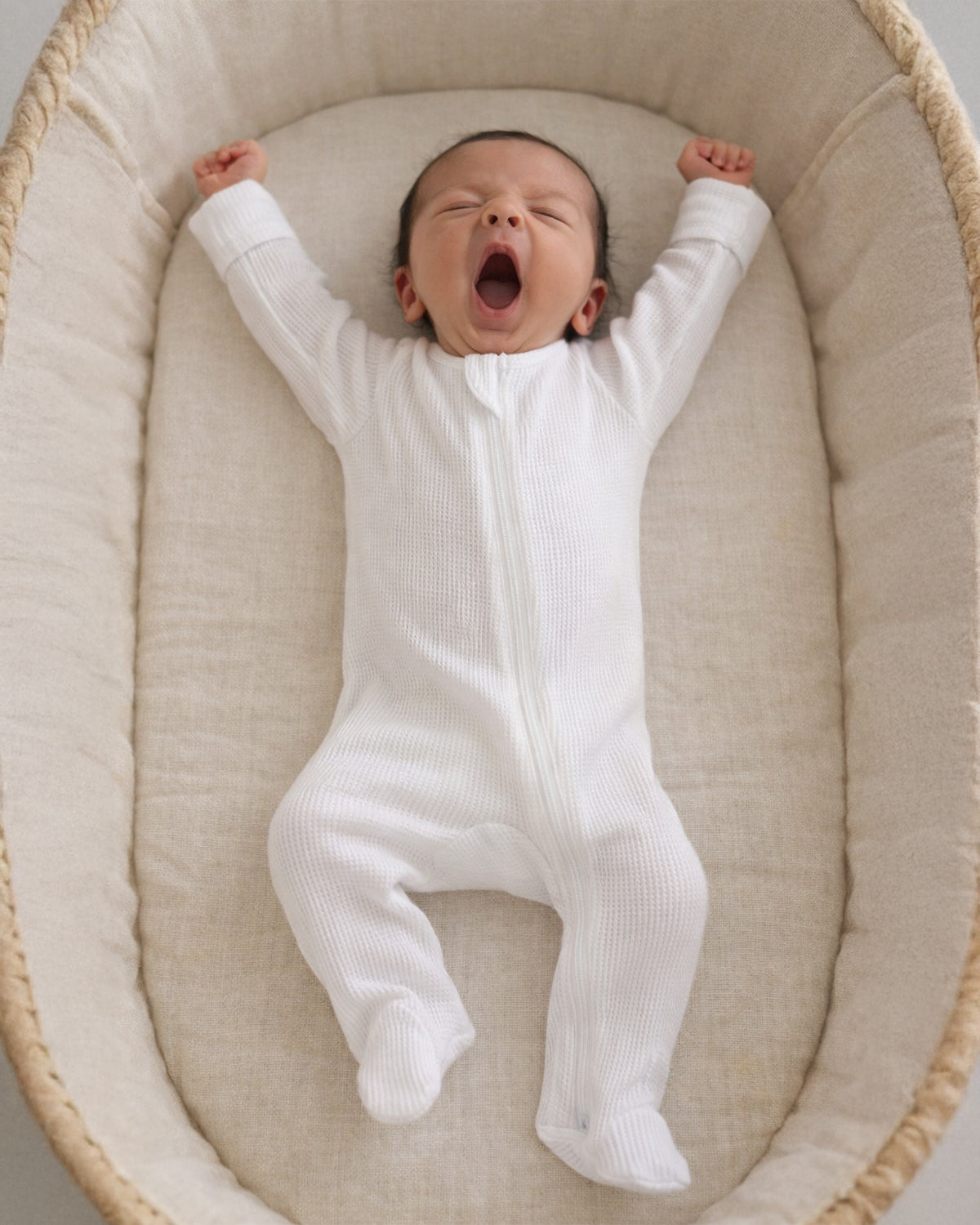 Baby in a white onesie yawning while lying in a cozy bassinet.