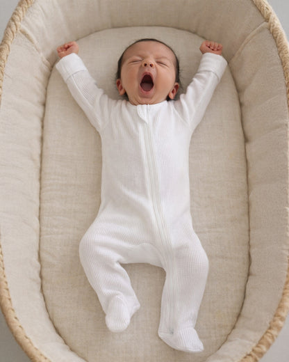 Baby in a white onesie yawning while lying in a cozy bassinet.