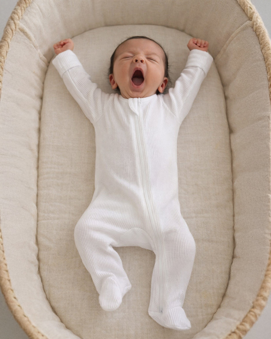 Baby in a white onesie yawning while lying in a cozy bassinet.