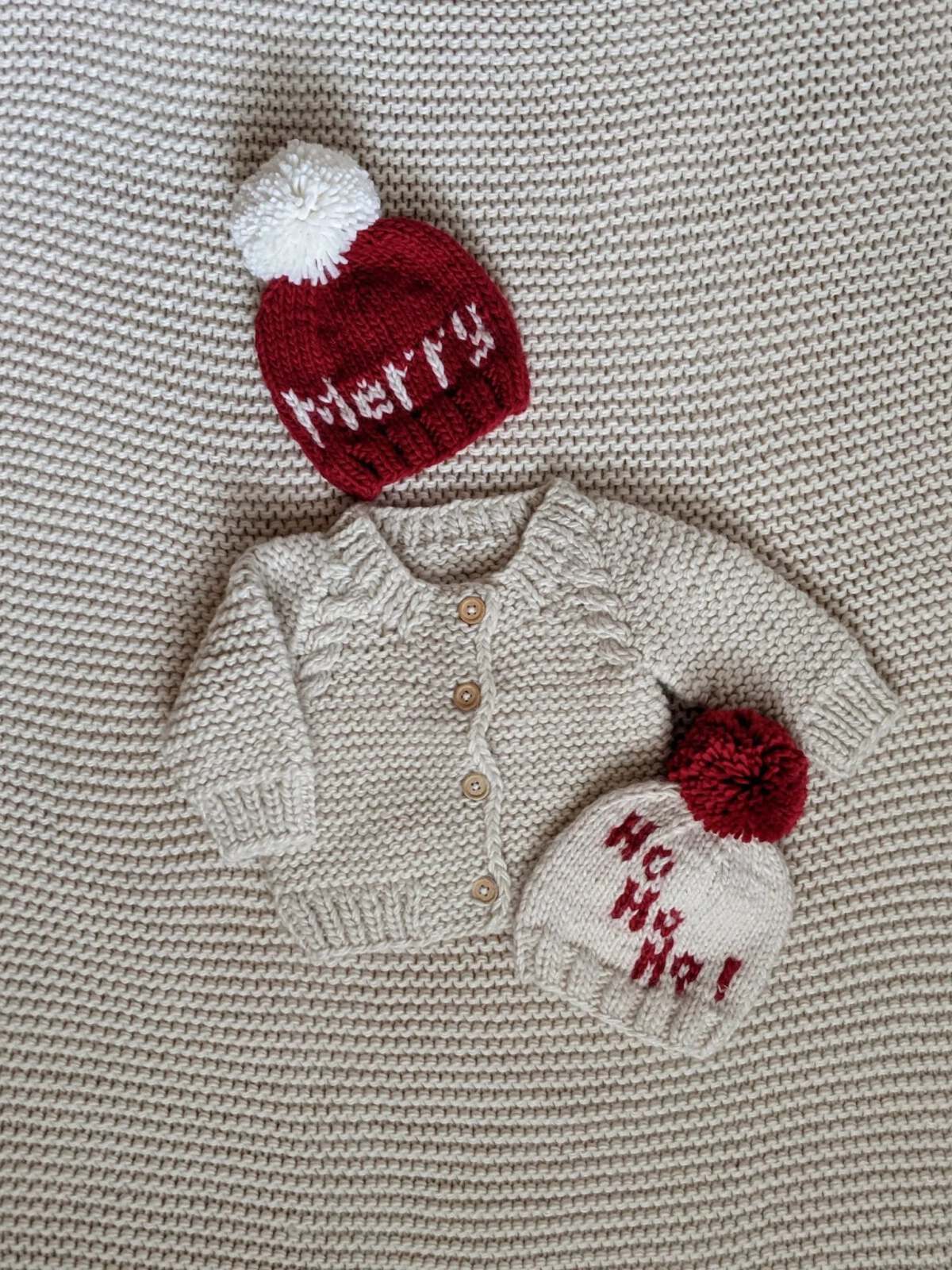 Baby cream cardigan with two holiday-themed knitted hats, one red with "Merry" and one white with "Ho Ho Ho!"