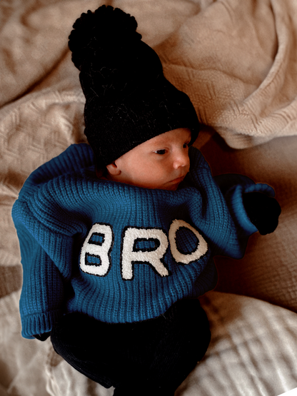 A baby in a blue sweater with "BRO" text, wearing a black beanie, rests on a blanket.