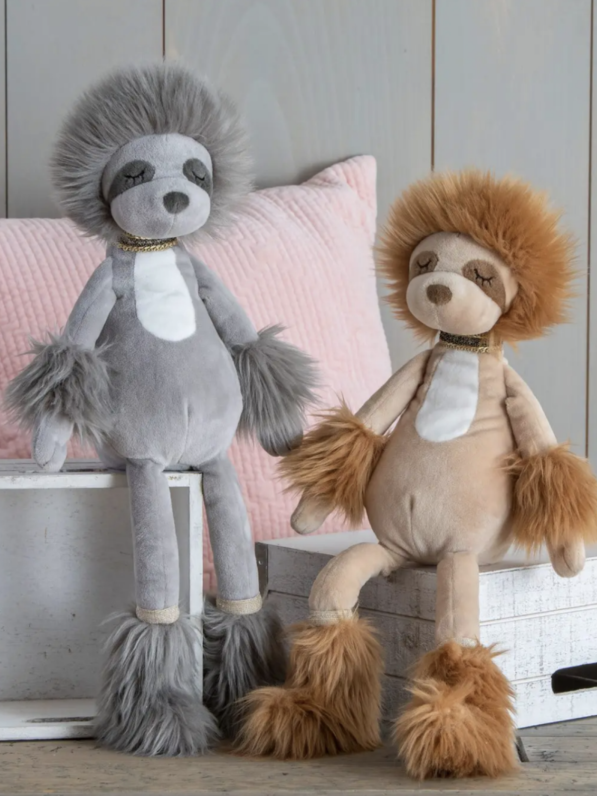 Two plush toy sloths with fluffy features, one gray and one tan, displayed on wooden boxes against a pastel background.