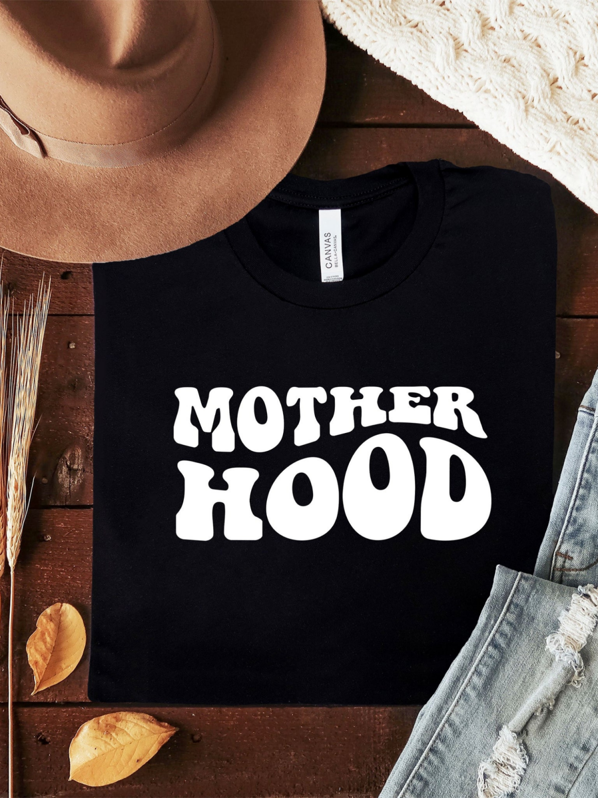 Black t-shirt with "MOTHER HOOD" in bold white letters, paired with a tan hat and jeans on a wooden background.