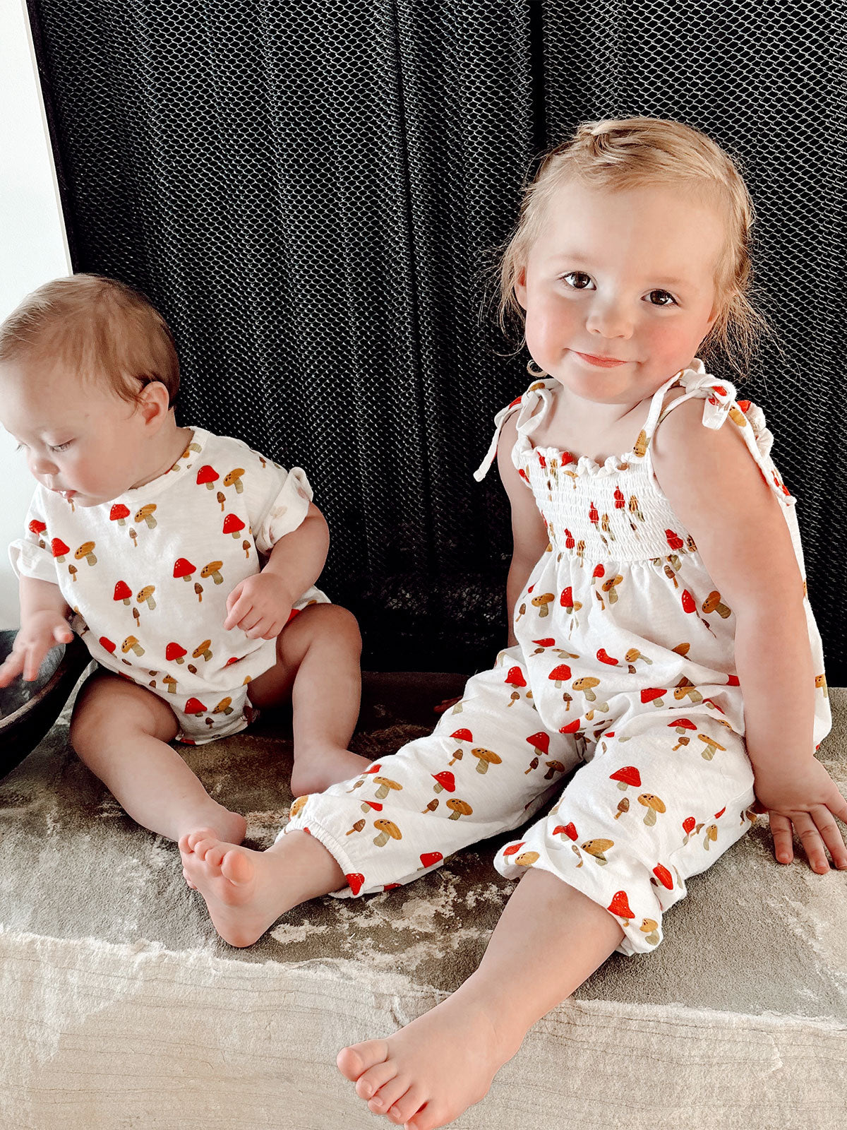 Two young children in matching outfits with mushroom prints sit on a rug near a black backdrop, smiling.