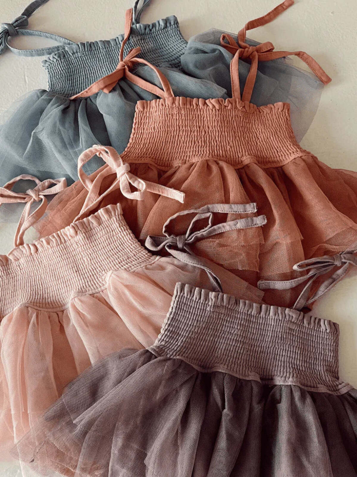 Four colorful children's dresses in soft tulle fabric, featuring smocked bodices and adjustable straps.