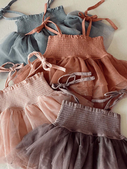 Four colorful children's dresses in soft tulle fabric, featuring smocked bodices and adjustable straps.