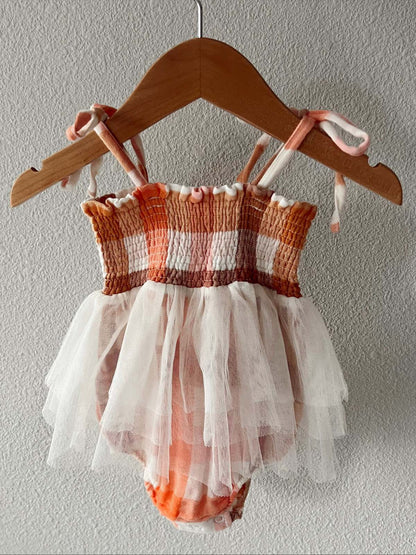 Peach and white checked toddler swimsuit with a tulle skirt, hanging on a wooden hanger against a textured wall.