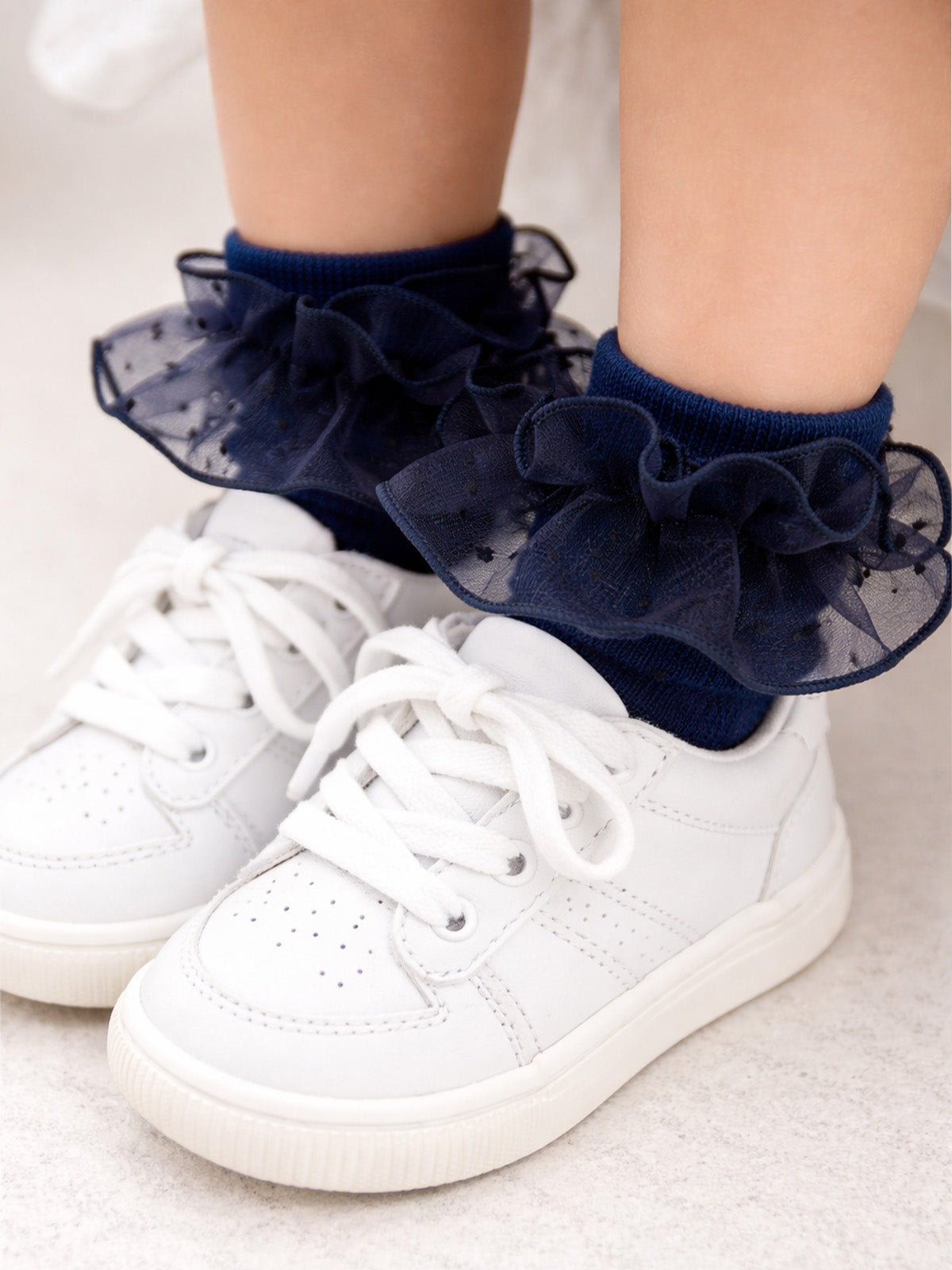 White sneakers with navy ruffled socks on a child's feet, set against a light background.