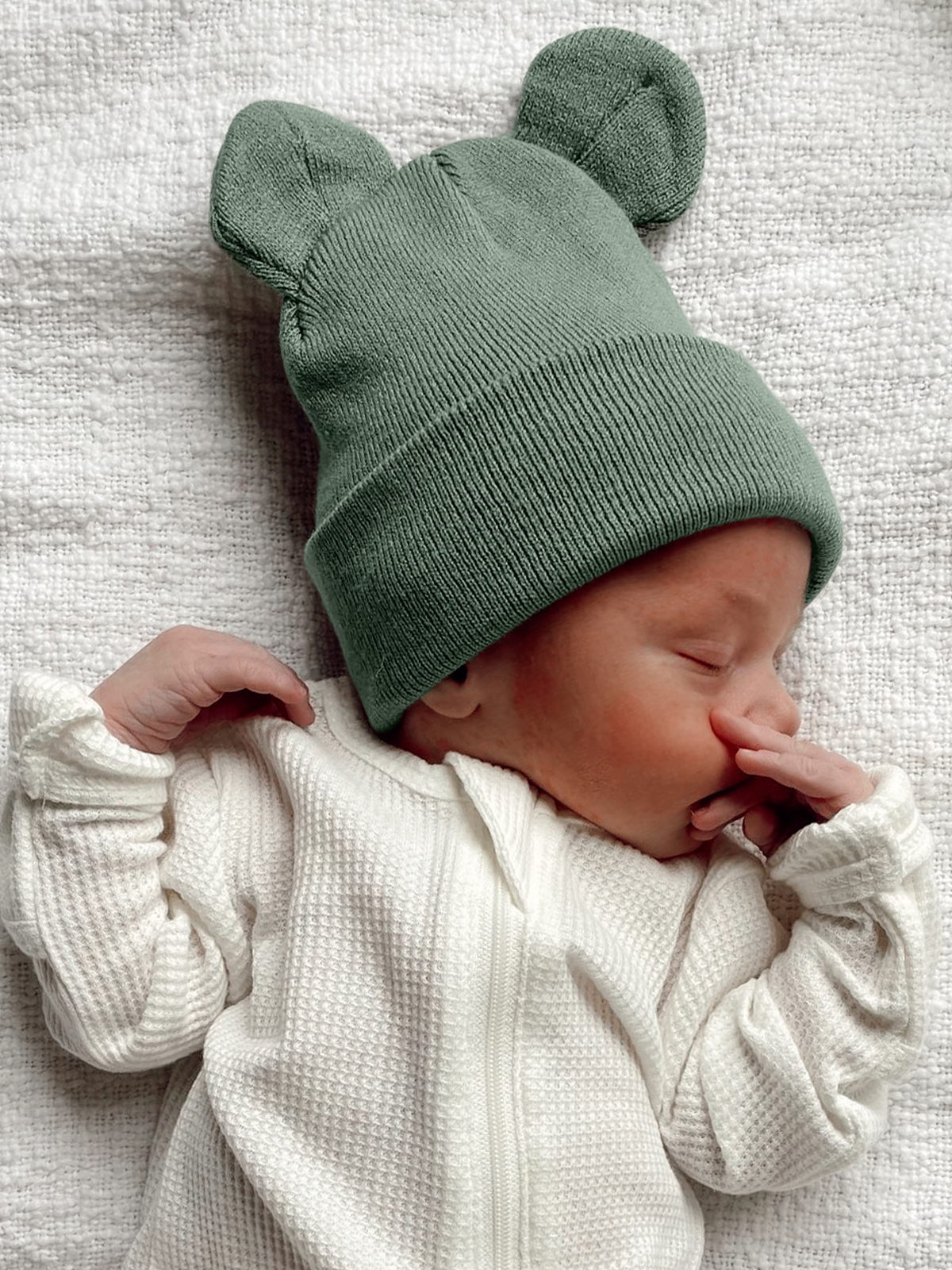 A sleeping baby in a green bear-ear beanie and white outfit, lying on a light textured blanket.