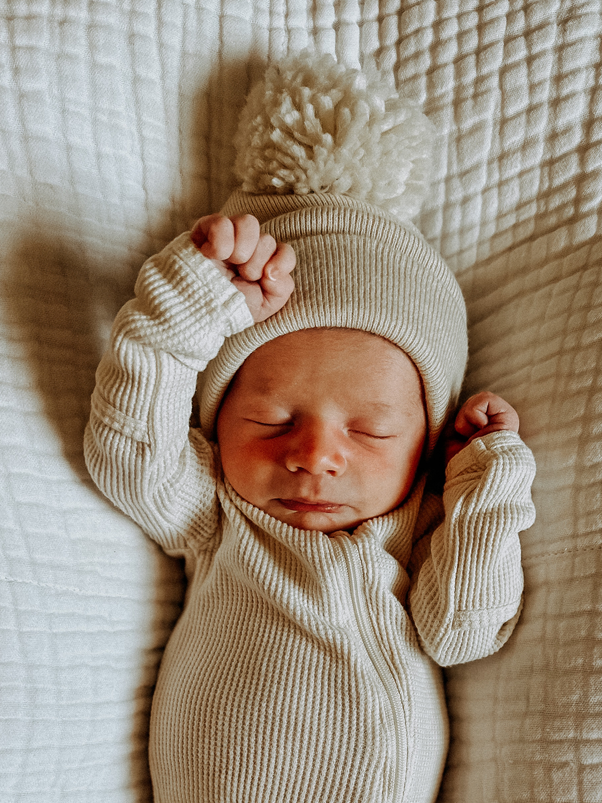 Newborn baby wearing a cream ribbed outfit and a fluffy pom-pom hat, sleeping on a quilted surface.