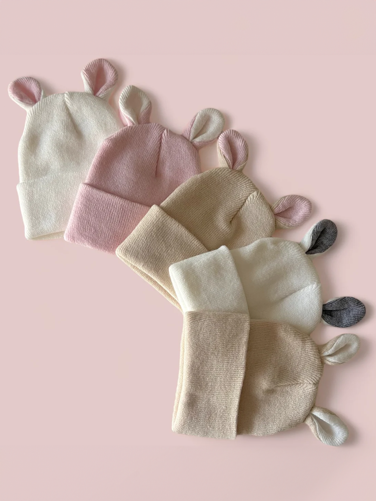 Four cute beanies with bunny ears in cream, pink, beige, and gray shades against a soft pink background.