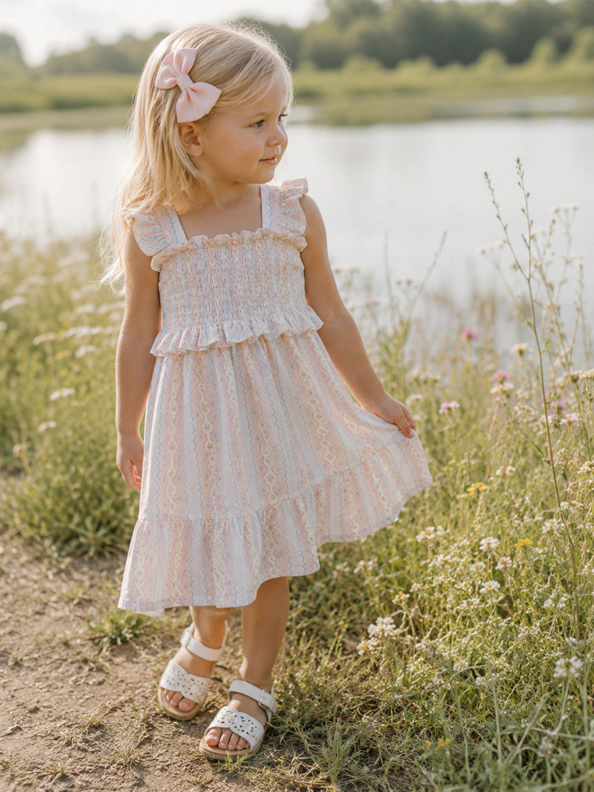 Girl in a pastel dress with a bow, walking through flowers by a calm pond.