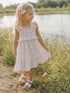 Girl in a pastel dress with a bow, walking through flowers by a calm pond.