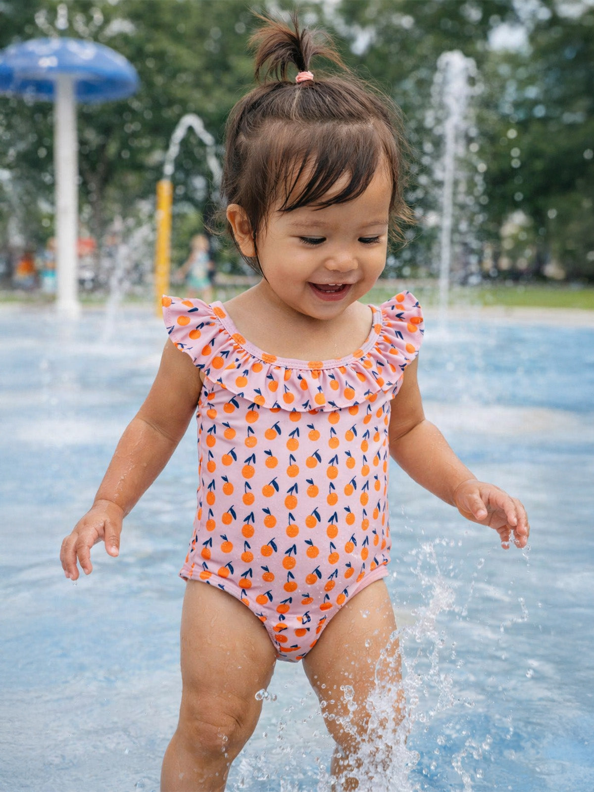 Toddler in a fruity-patterned swimsuit joyfully splashes in a fountain at a park on a sunny day.