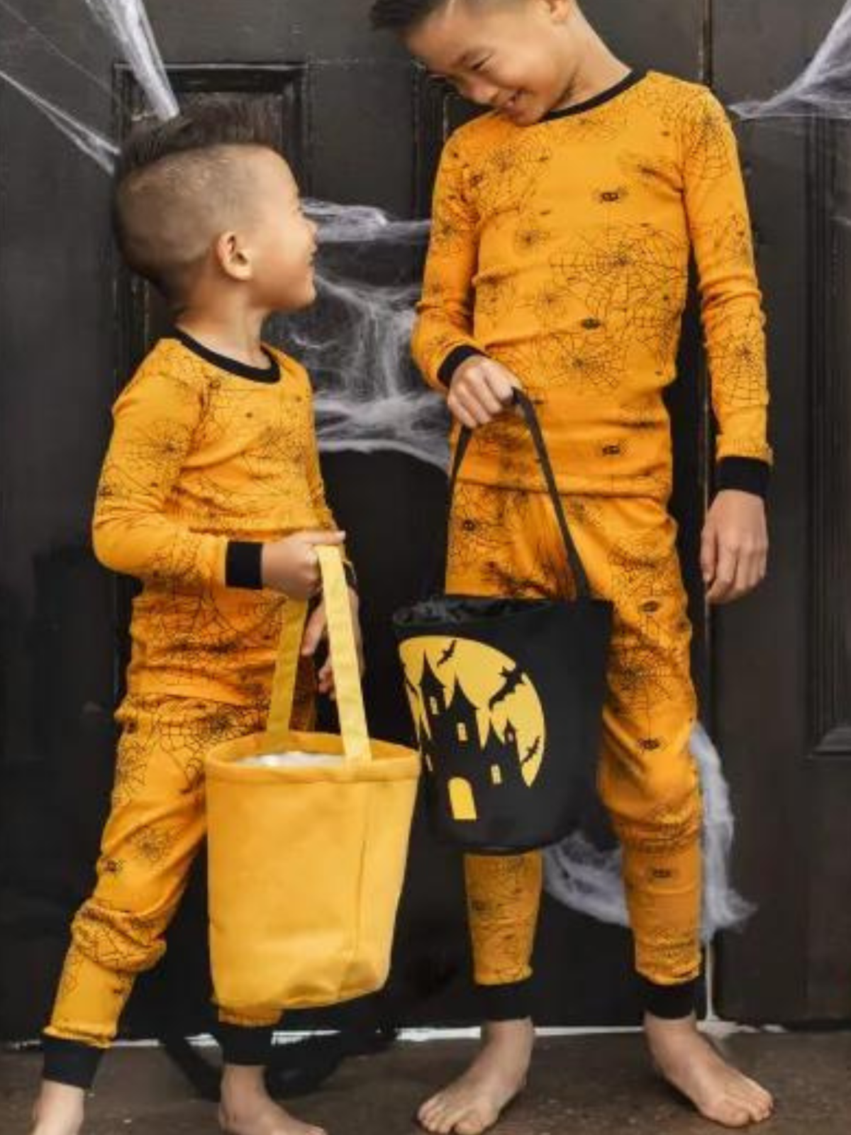 Two boys in orange spider-print pajamas hold Halloween baskets in front of a door decorated with spider webs.