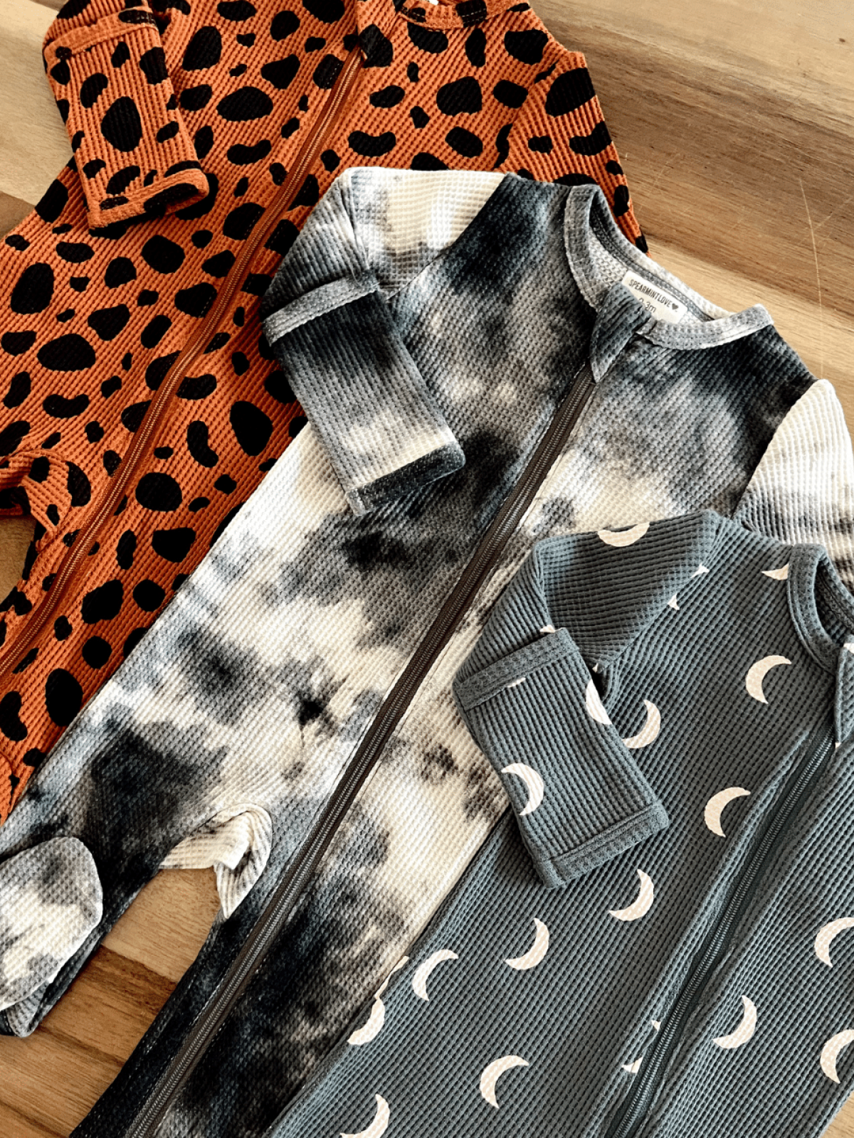 Three cozy baby onesies in animal print, tie-dye, and crescent moon designs, displayed on a wooden surface.