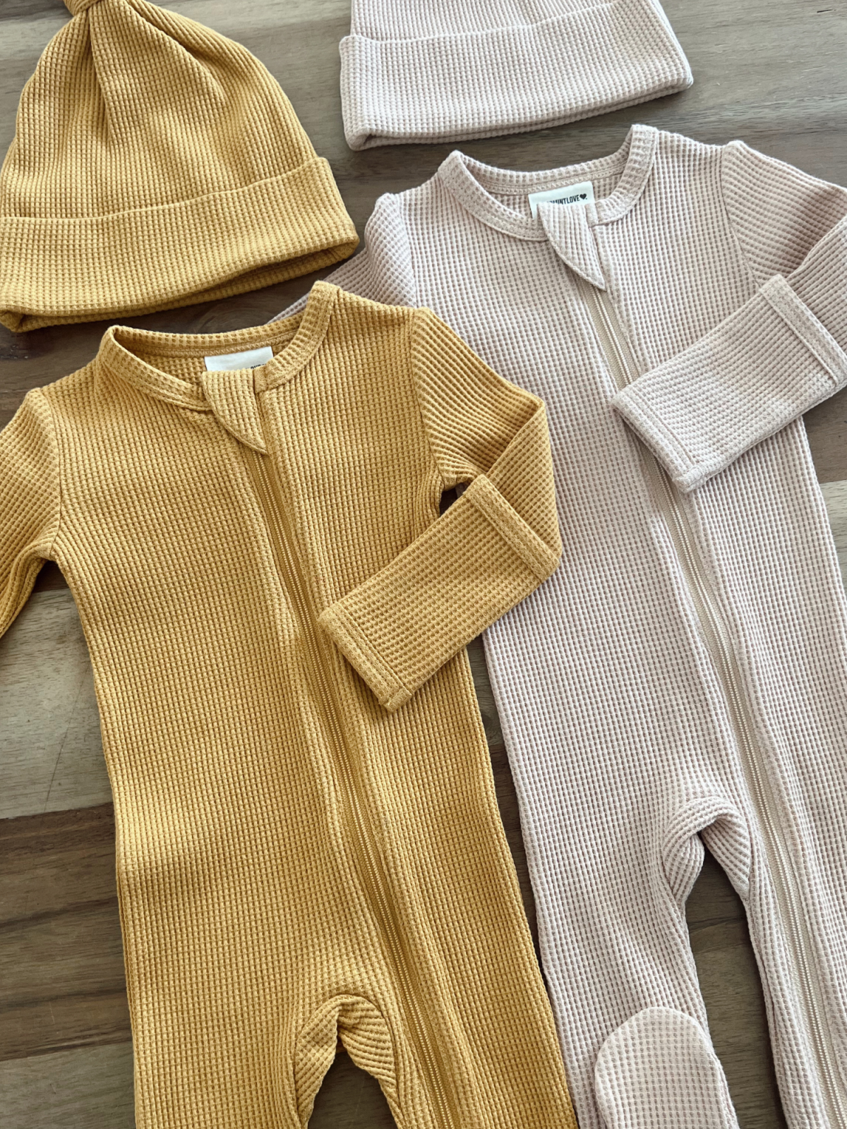 Two infant bodysuits in mustard and light pink, paired with matching beanies, arranged on a wooden surface.