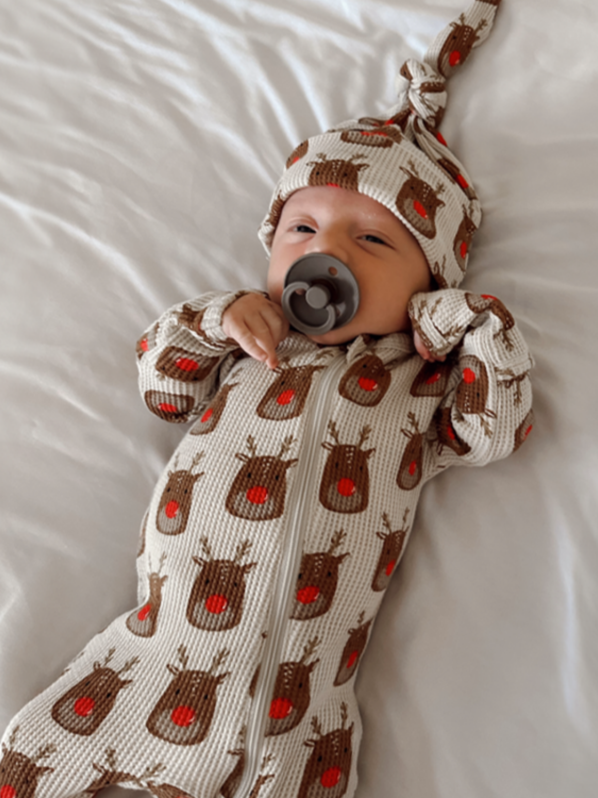 Infant in a cozy reindeer-patterned outfit with a pacifier, lying on a soft white blanket.