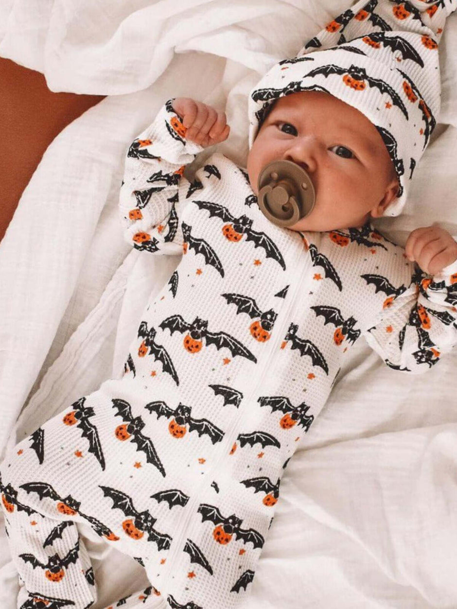 Baby in a cozy Halloween-themed outfit with bats and pumpkins, lying on a blanket.