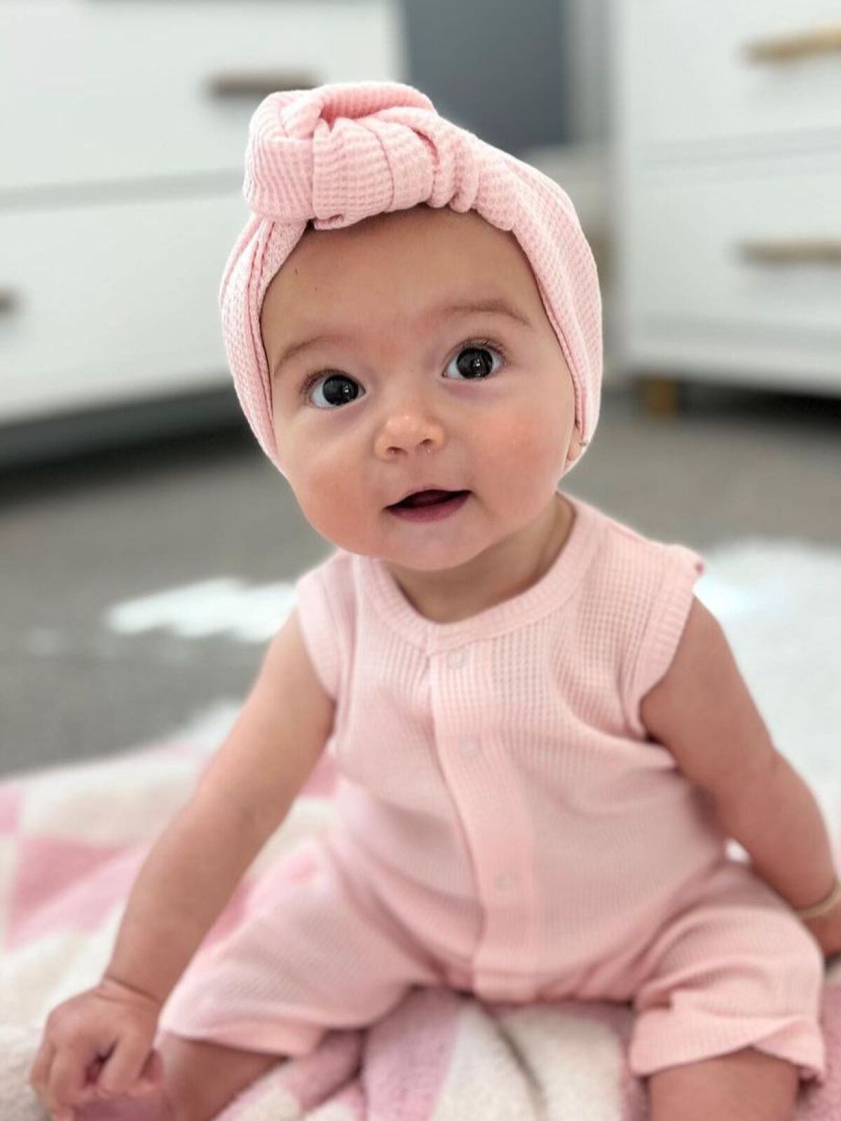 Smiling baby girl in a pink outfit and headband, seated on a blanket in a bright room.