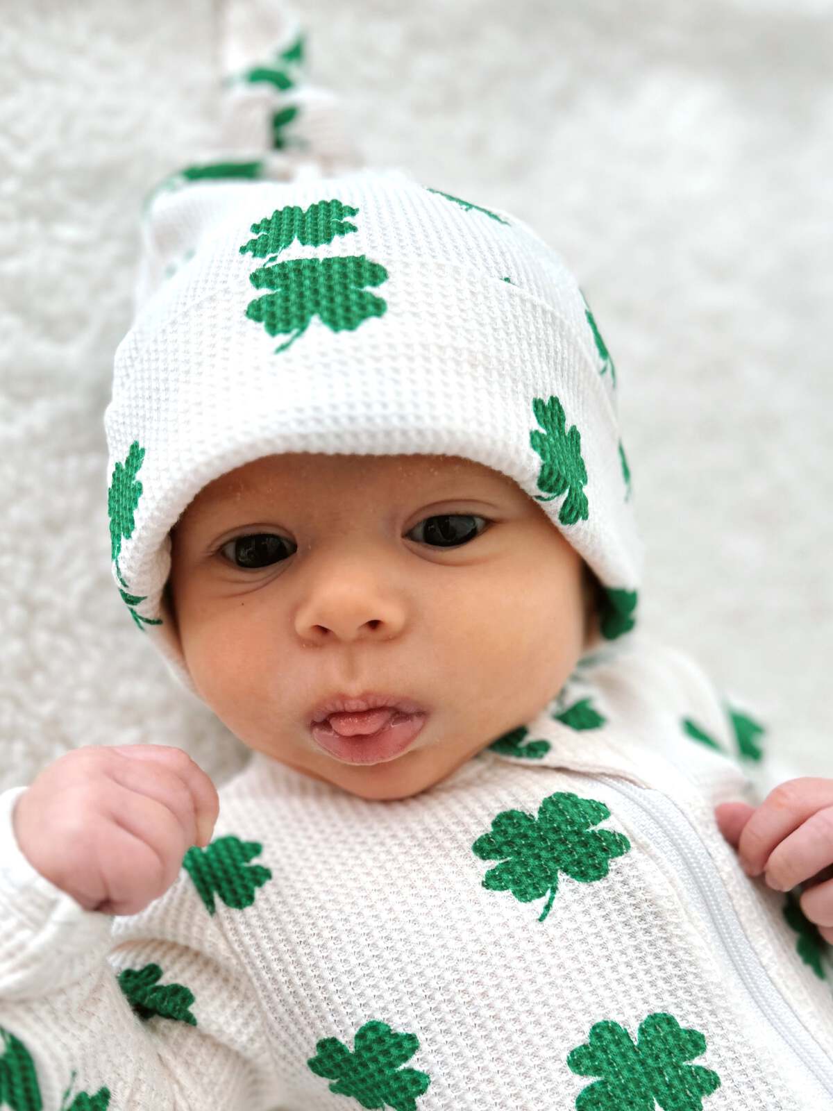 Baby wearing a white outfit with green shamrocks, looking curiously at the camera with a playful expression.