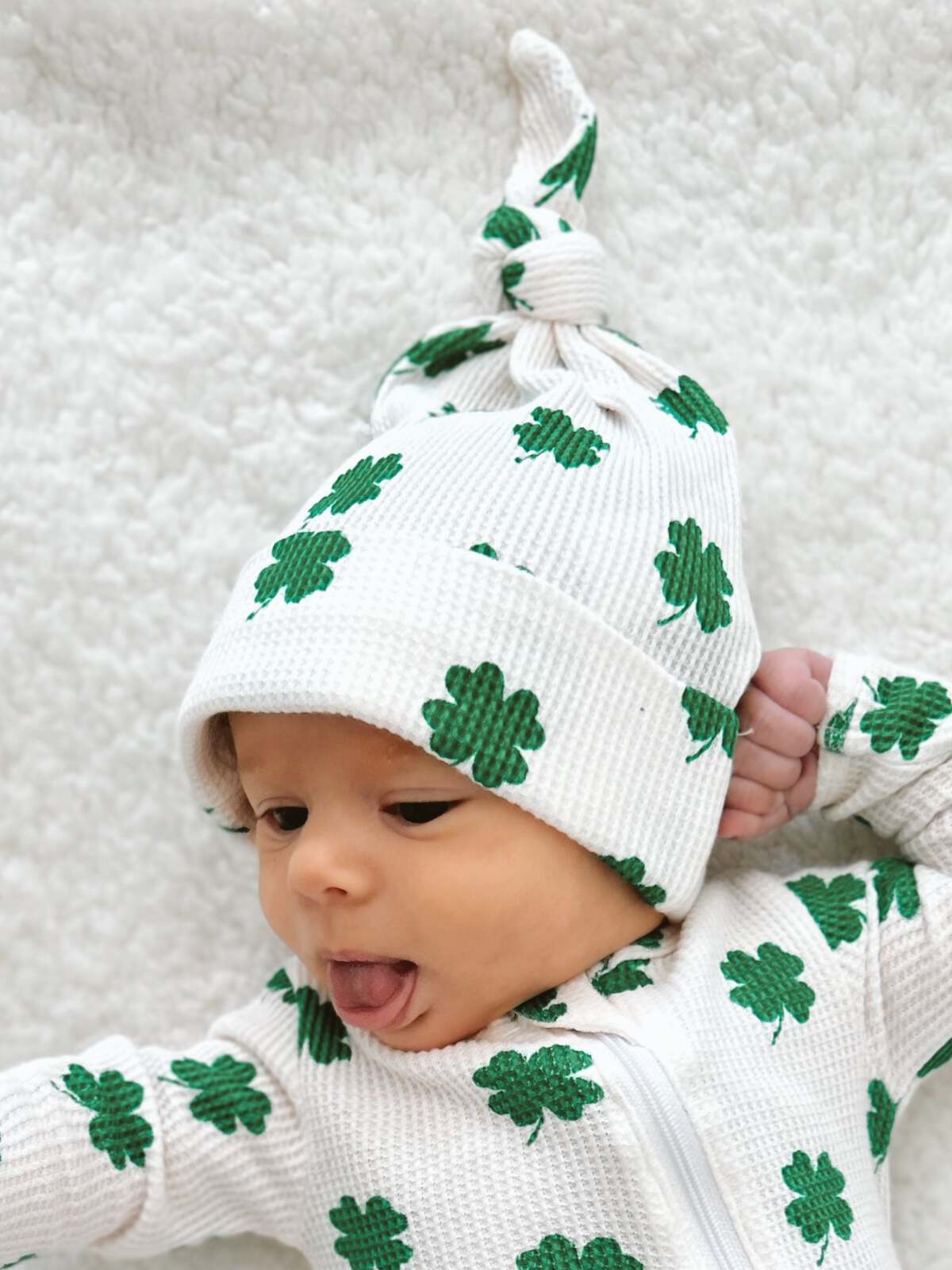 Baby wearing a white outfit with green clovers, lying on a soft surface, sticking out their tongue playfully.