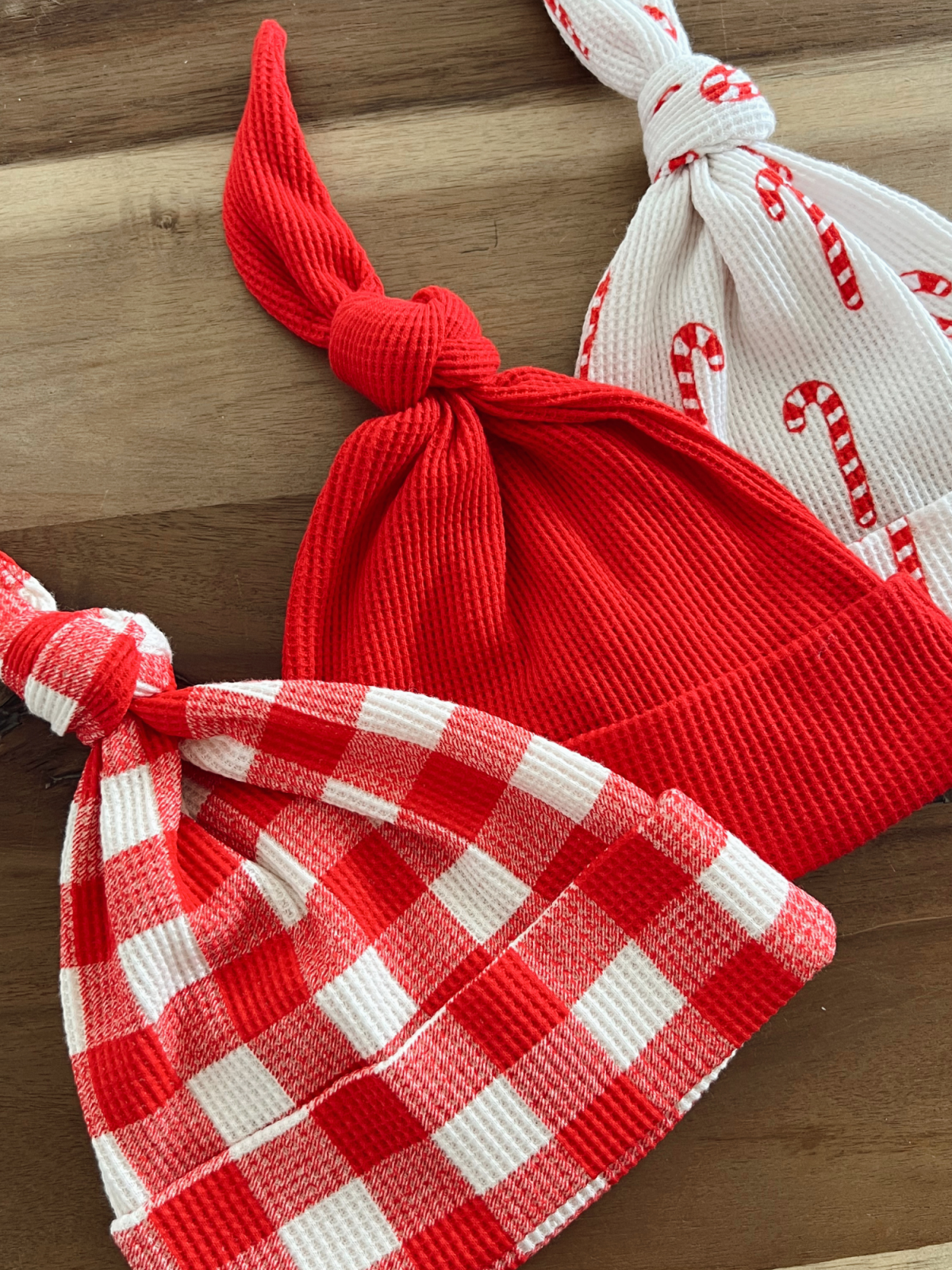 Three festive hats: a solid red, a red and white checkered, and a white with candy cane pattern on a wooden surface.
