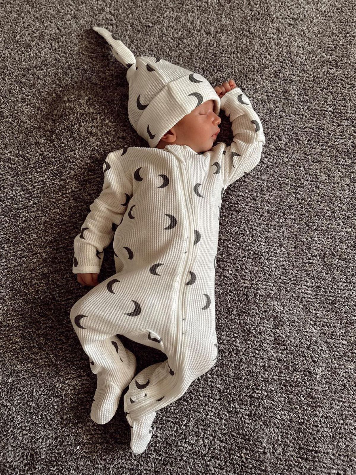 Newborn sleeping on a gray rug, wearing a white outfit with crescent moon patterns and a matching hat.