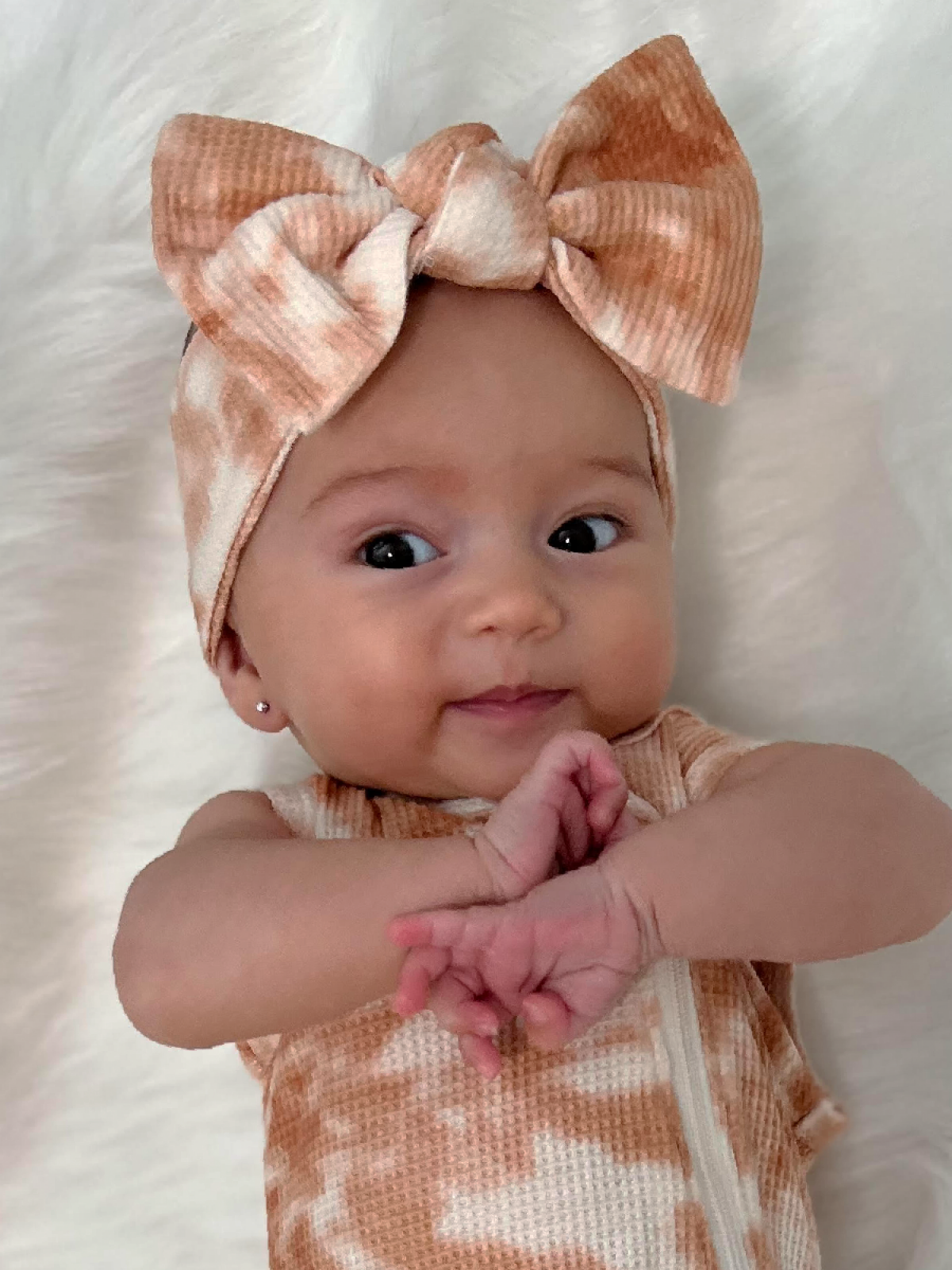 Smiling baby wearing a peach patterned outfit and large bow headband, lying on a soft white surface.