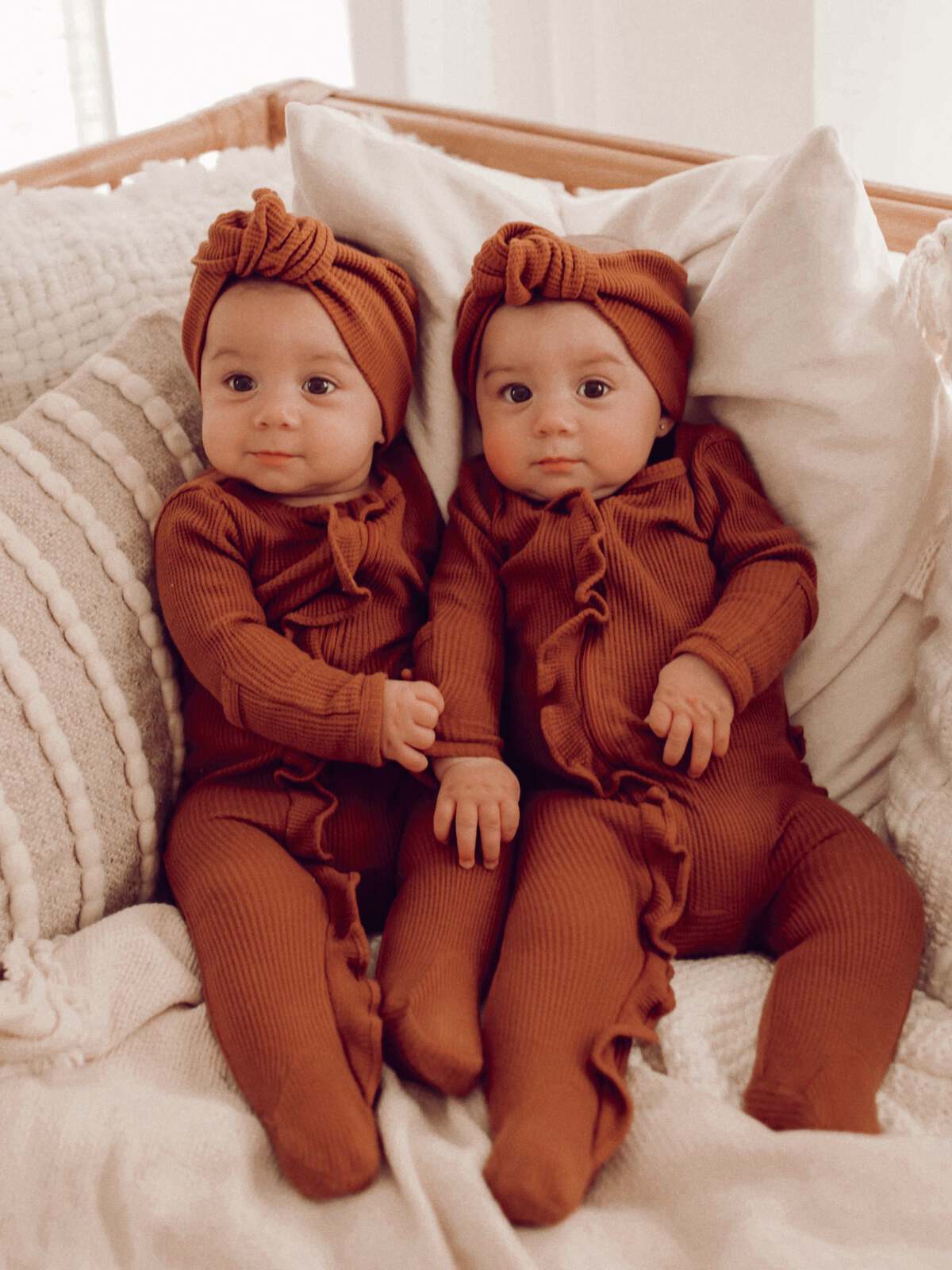 Two identical babies in brown outfits and headbands are sitting together on a cozy blanket.