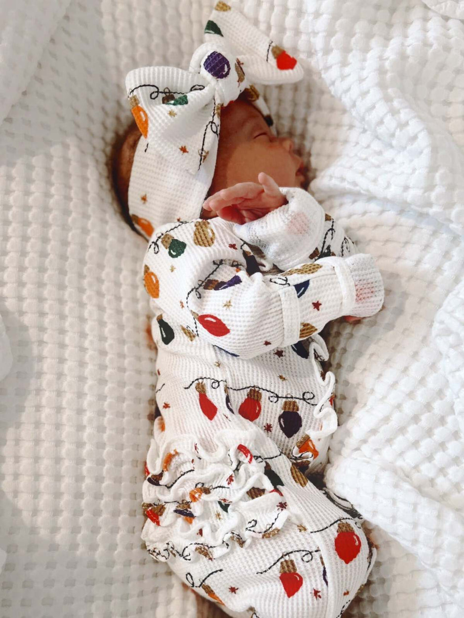 Newborn in festive pajamas with a bow, lying on a cozy white blanket.